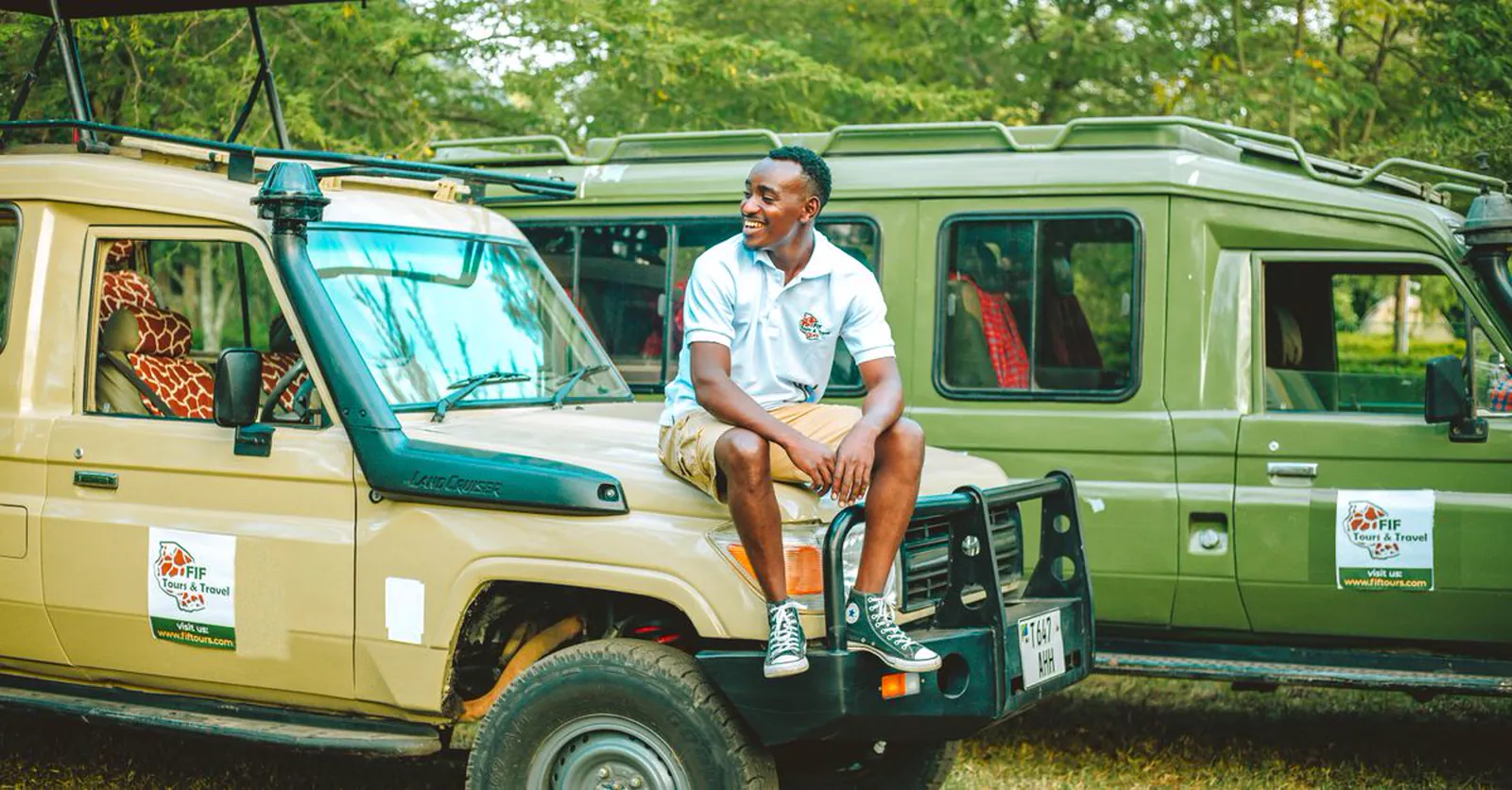 A smiling safari guide seated on an open safari vehicle with a giraffe behind him.