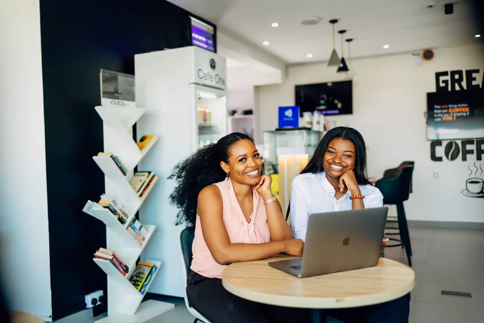 Two smiling women sit at a round table with a laptop in a bright Lagos cafe.