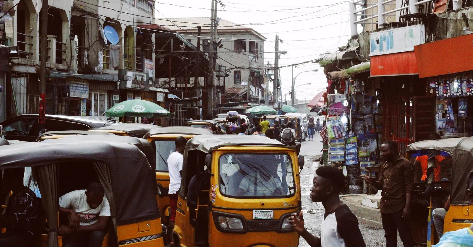 A busy Nigerian street lined with small shops and signboards, with yellow keke tricycles queued along the road.