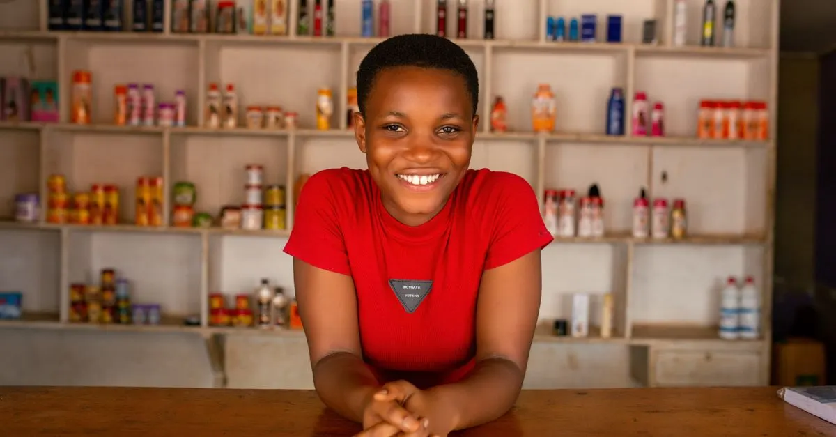 A smiling shopkeeper in a red T-shirt leans on a wooden counter in front of shelves of toiletries and household goods.