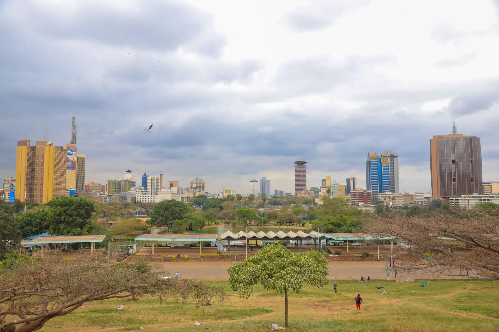 The Nairobi city skyline seen across Uhuru Park under a cloudy sky, with office towers behind the trees.
